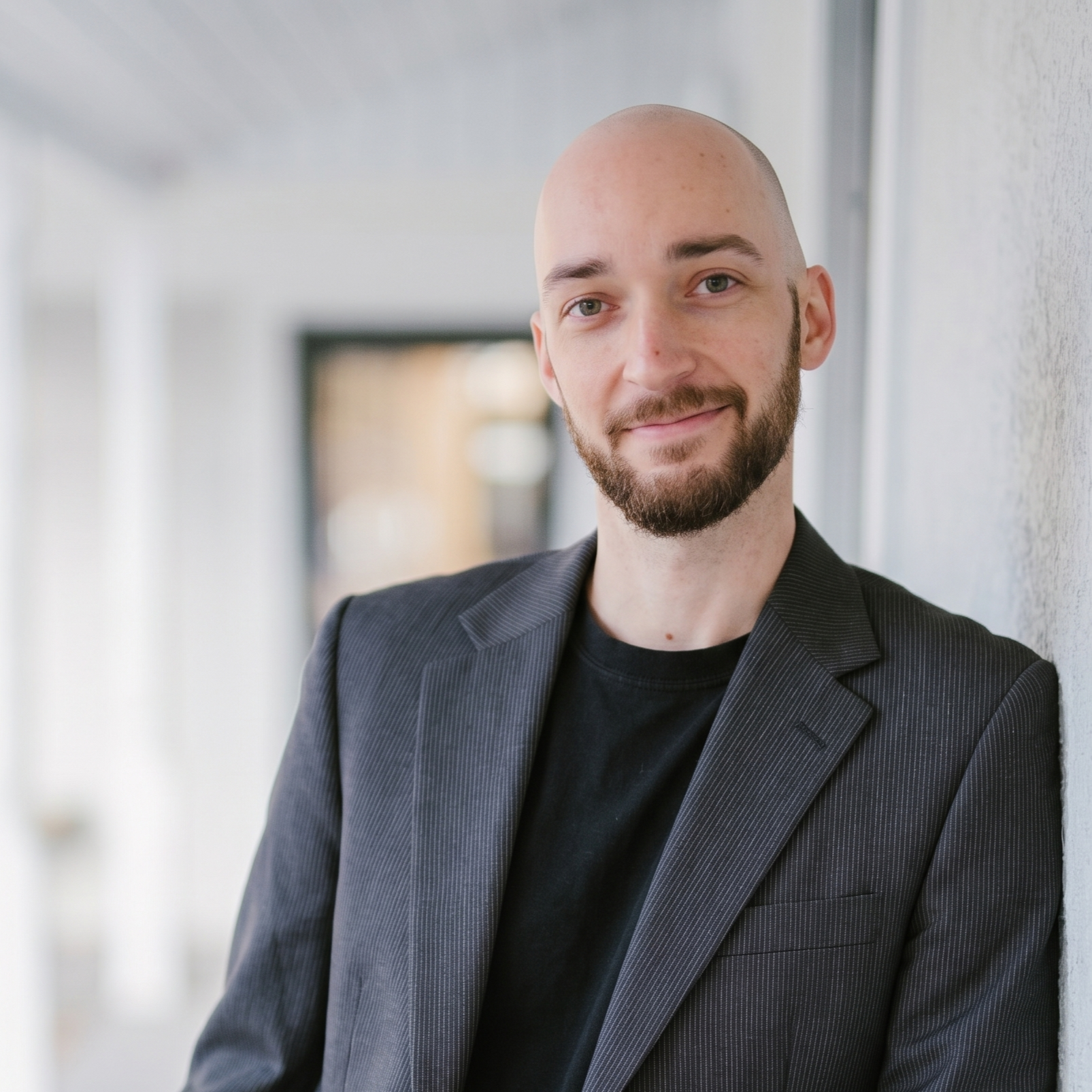 kevin picture2 Smiling man with a shaved head and beard wearing a dark blazer stands in a bright corridor.